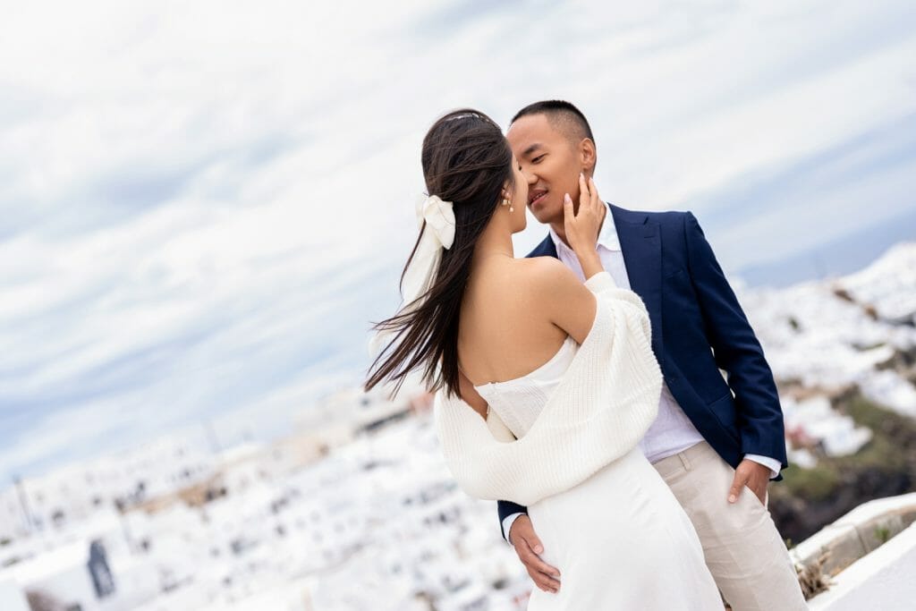 couple sharing a close, warm hug, expressing affection and happiness, with Santorini’s whitewashed buildings softly blurred in the background.