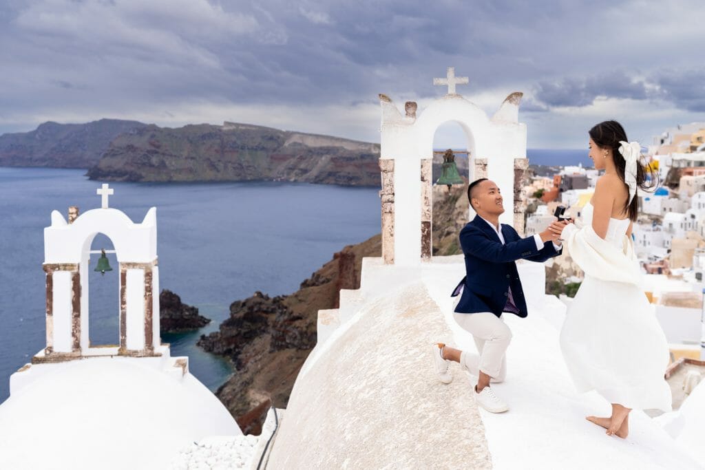A man on one knee holding out a ring to his surprised girlfriend, both smiling warmly with the picturesque Santorini cliffs and blue sky behind them.