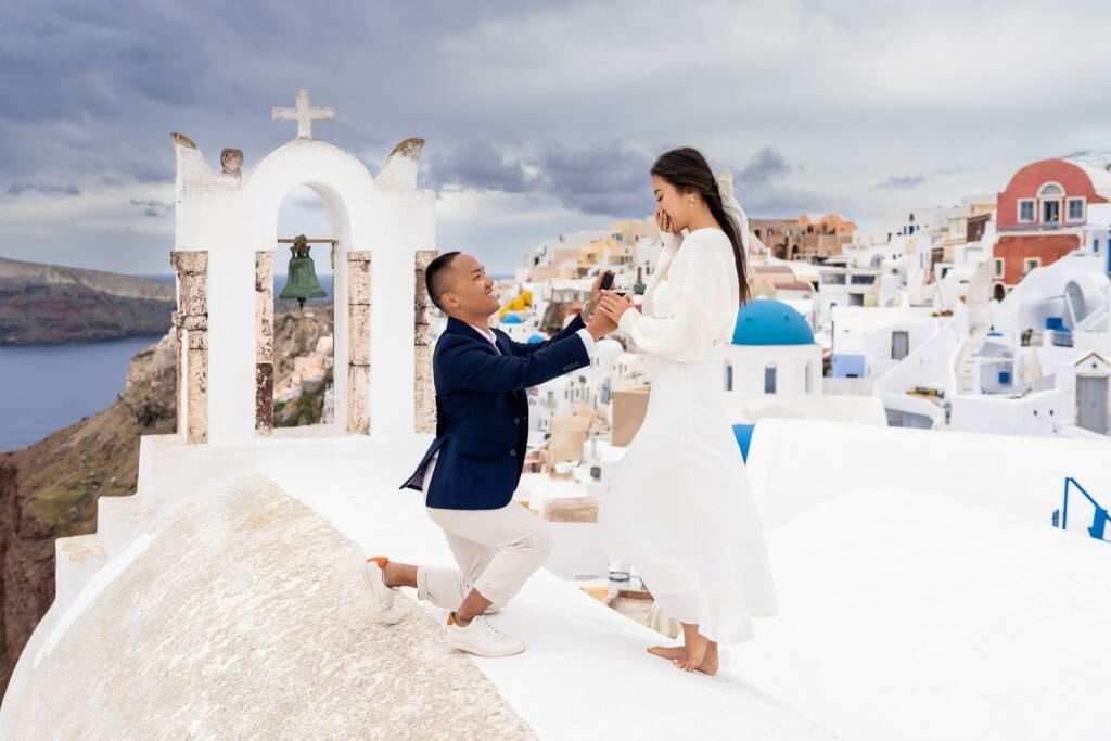 A man kneeling to propose with a ring in hand to his girlfriend, who is standing tall, against the stunning backdrop of Santorini’s white buildings and blue sea.