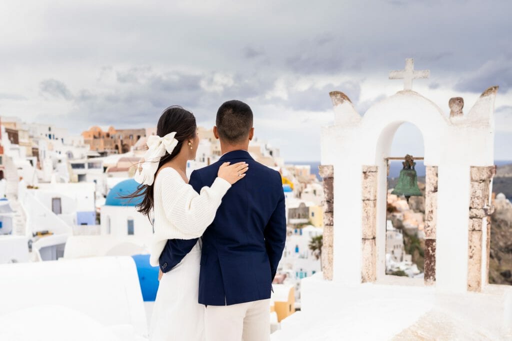 A couple enjoying the view of the iconic Santorini church bell, framed by white architecture and a bright blue sky.