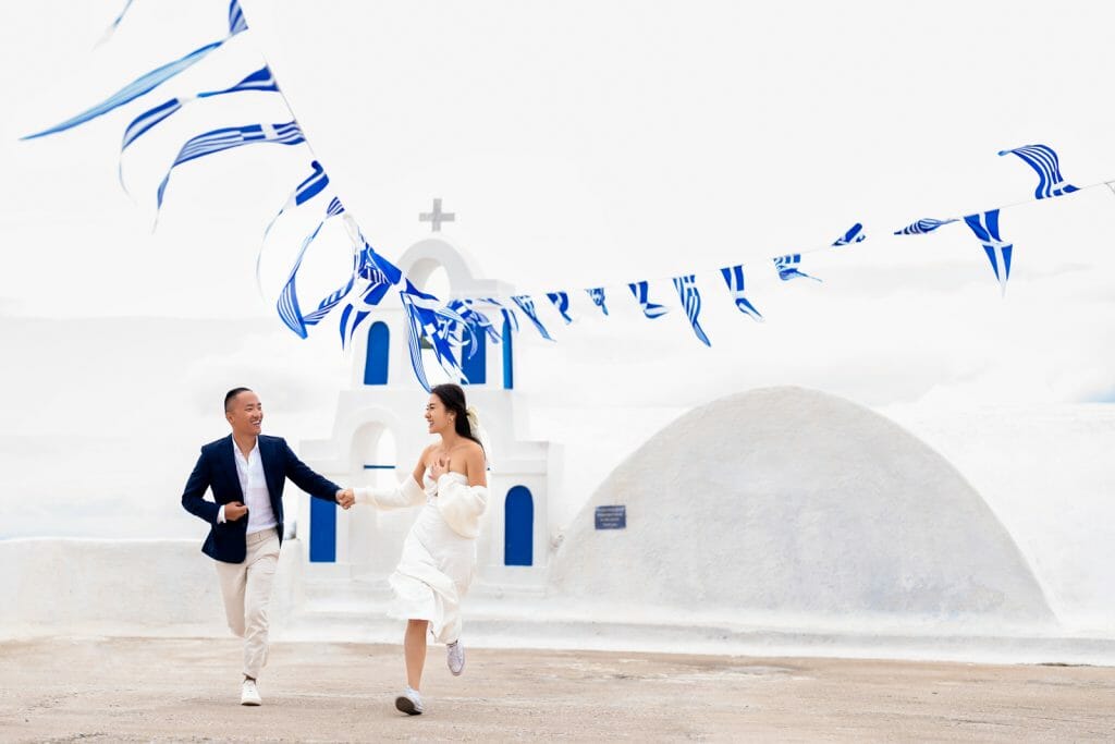 A happily married couple running joyfully together in Santorini with a white church and blue dome in the background, surrounded by the island’s iconic bright architecture.