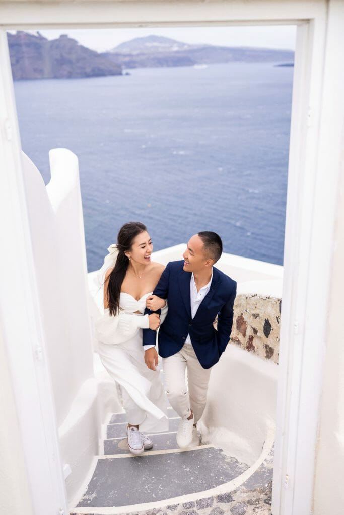 A happy couple walking up the white stone stairs in Santorini, surrounded by iconic white buildings and blue domes, enjoying a romantic moment under the bright Mediterranean sun.