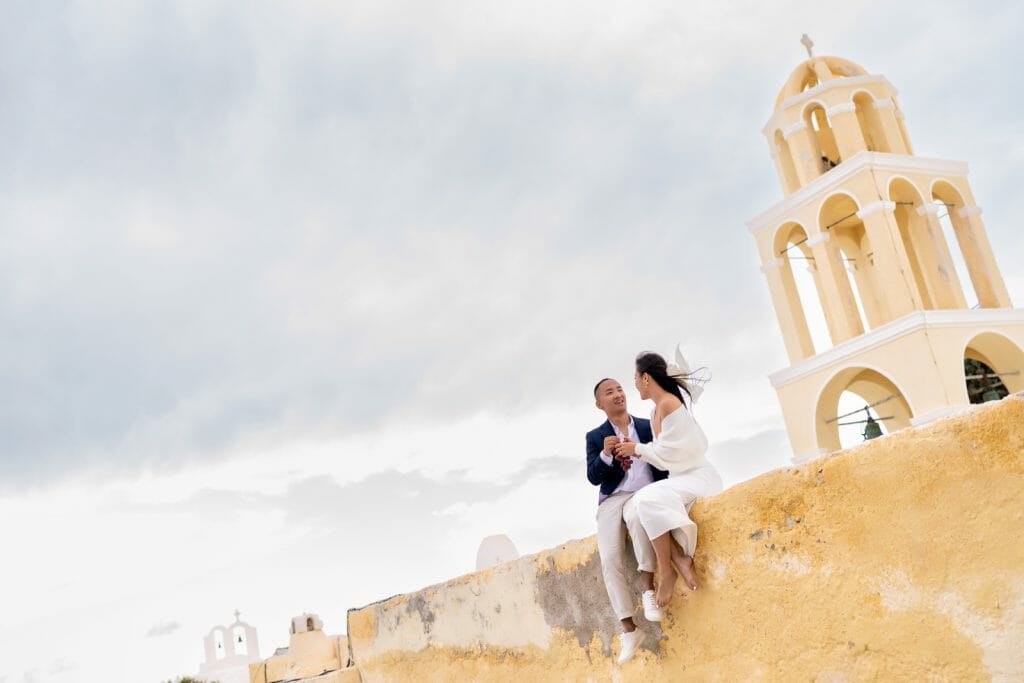 A couple posing for photos in front of the stunning Santorini landscape, with whitewashed buildings, blue domes, and the sparkling Aegean Sea in the background.