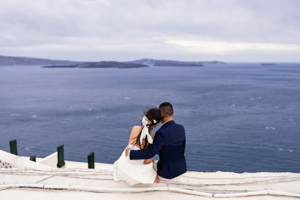 A couple standing with their backs to the camera, embracing in front of the sea during a romantic Santorini photoshoot, with the blue Aegean Sea and whitewashed architecture in the background.