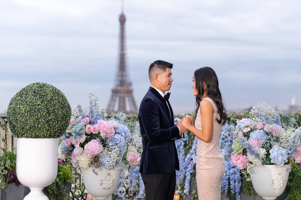 Gentleman proposes on Peninsula Paris rooftop with Eiffel Tower view, flowers, and candlelight. He gazes into her eyes.