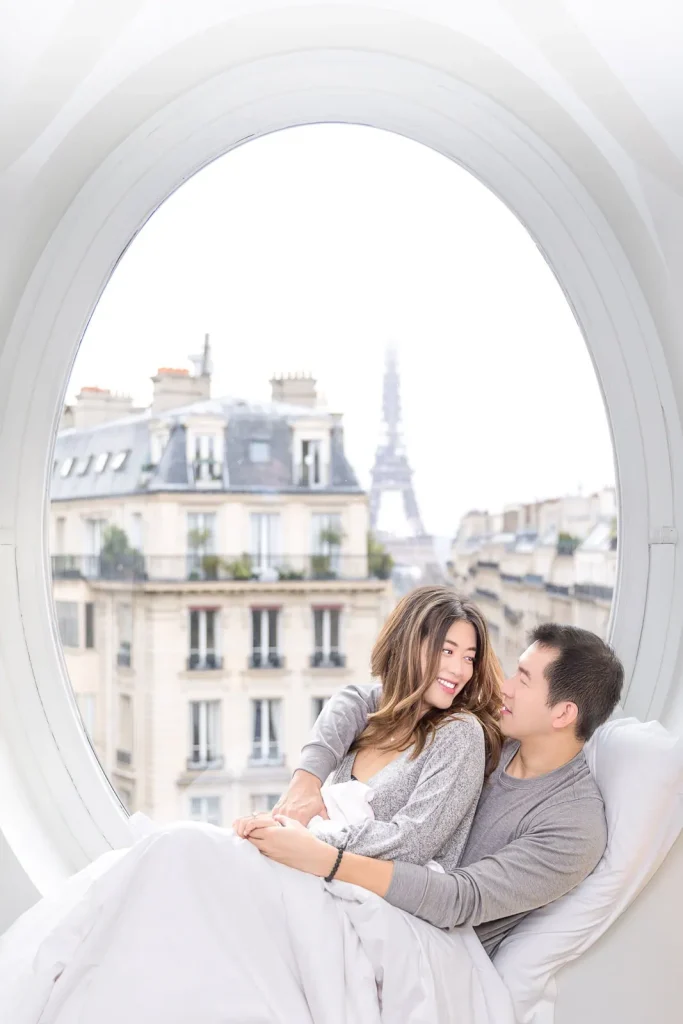 Playful couple cuddling in bed during indoor Paris engagement photo shoot with Eiffel Tower visible through oval window at Le Metropolitan Hotel
