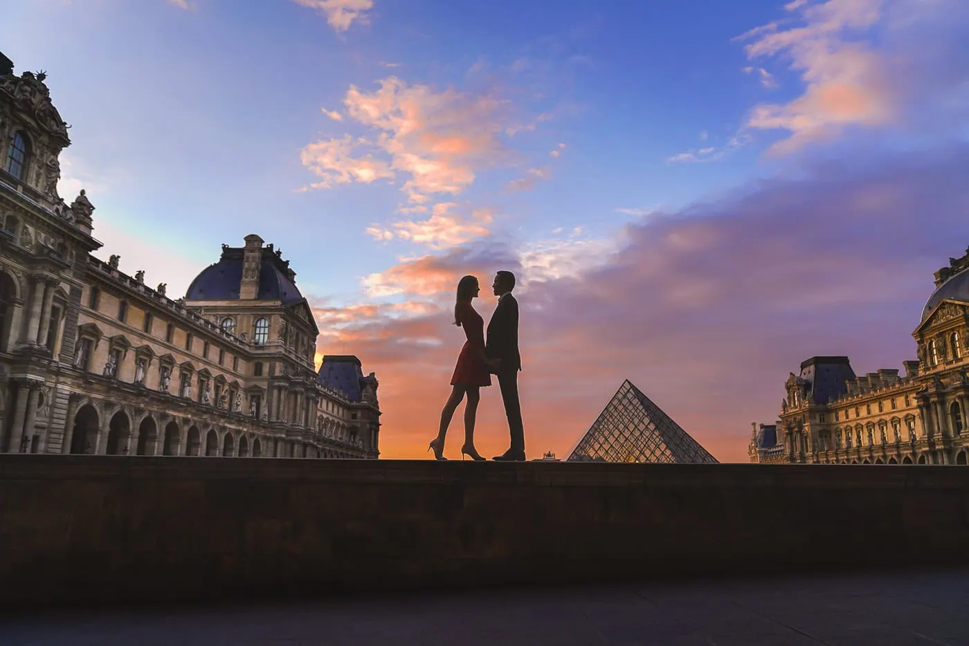 Couple in silhouette with woman in red dress against Louvre pyramid during Blue Hour, photographed from rear courtyard for crowd-free composition.