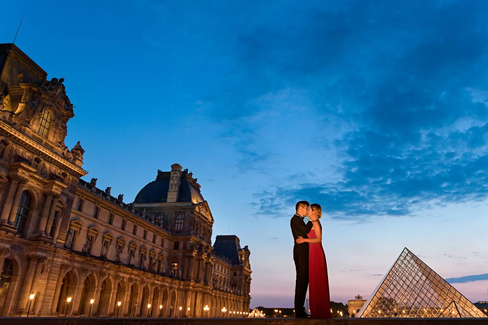 Engaged couple kissing at Louvre during blue hour with illuminated glass pyramid and romantic lighting