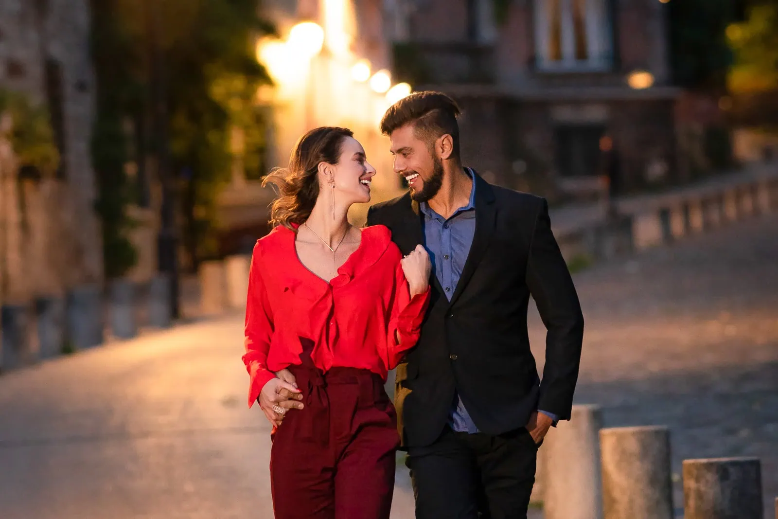 Una pareja comprometida pasa por La Maison Rose en Montmartre por la noche con un abrazo íntimo y contacto visual.