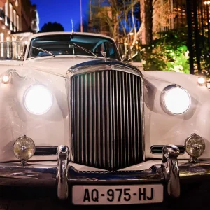 Белый старинный автомобиль с включенными фарами возле отеля Shangri-La Paris в синий час, готовый к трансферу с шофером.