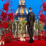 Elegant gentleman in a tuxedo standing on the Shangri-La rooftop terrace surrounded by red roses and a red carpet, looking at the illuminated Eiffel Tower against a vivid blue sky