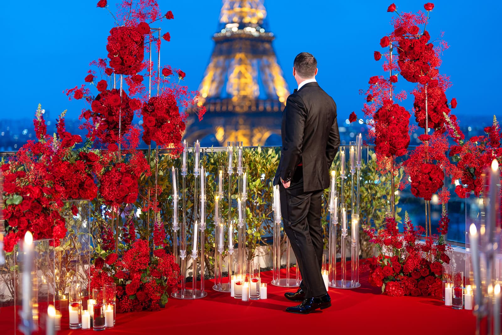 Elegant gentleman in a tuxedo standing on the Shangri-La rooftop terrace surrounded by red roses and a red carpet, looking at the illuminated Eiffel Tower against a vivid blue sky