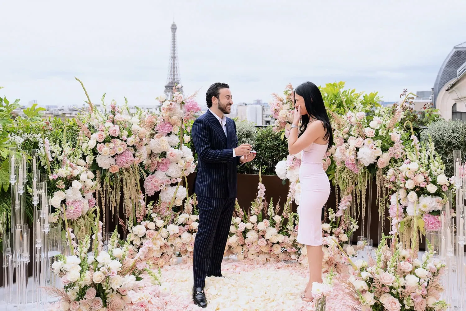 Мужчина дарит бриллиантовое кольцо на террасе ресторана The Peninsula Paris Secret Table, в то время как его партнерша прикрывает рот слезами; пол из роз в форме сердца и Эйфелева башня позади.