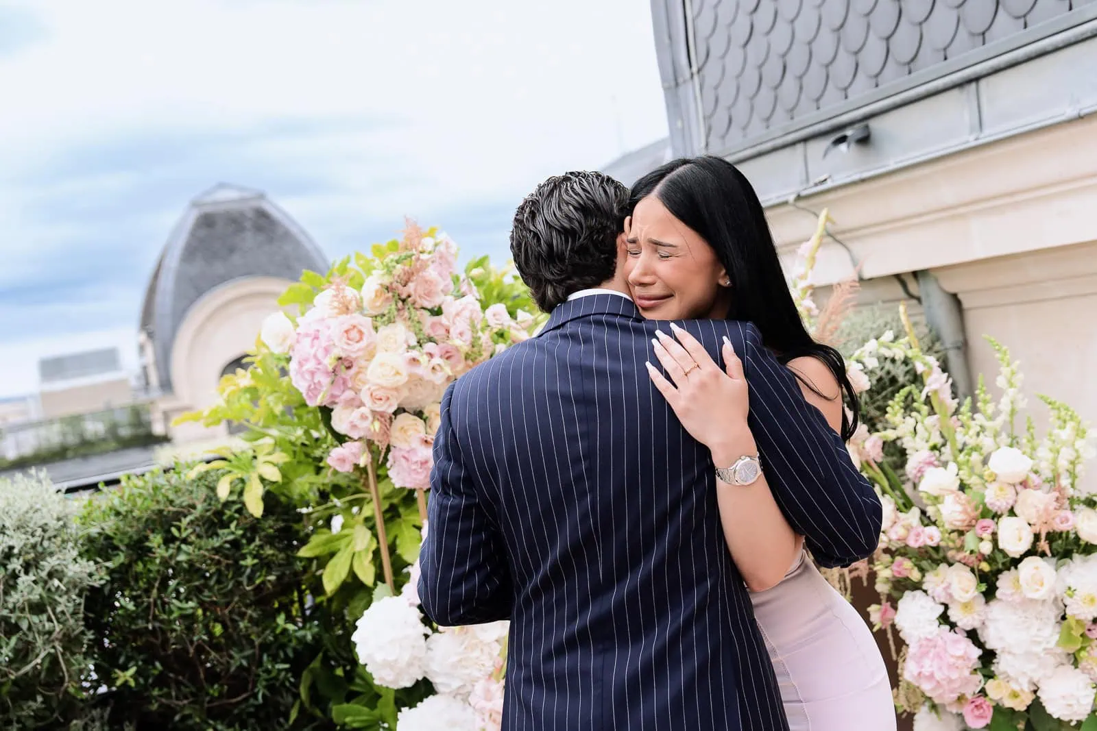Недавно помолвленная партнерша, с глазами, полными слез, обнимает своего жениха на террасе The Peninsula Paris Secret Table рядом с композициями из белых и розовых роз.