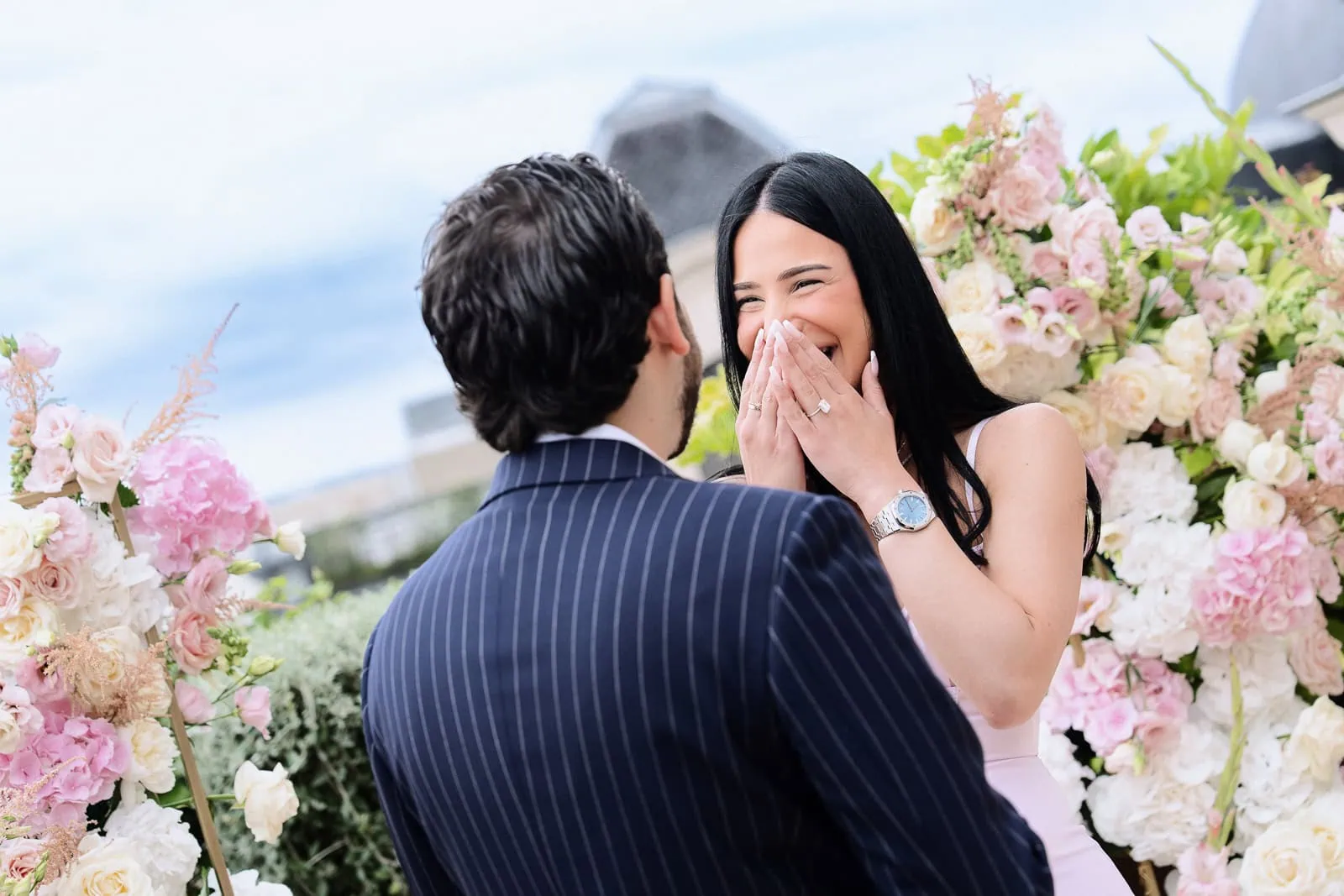 Невеста, переполненная эмоциями, закрыла лицо, делая романтическое предложение на Peninsula Paris Secret Table с лепестками роз и видом на Эйфелеву башню