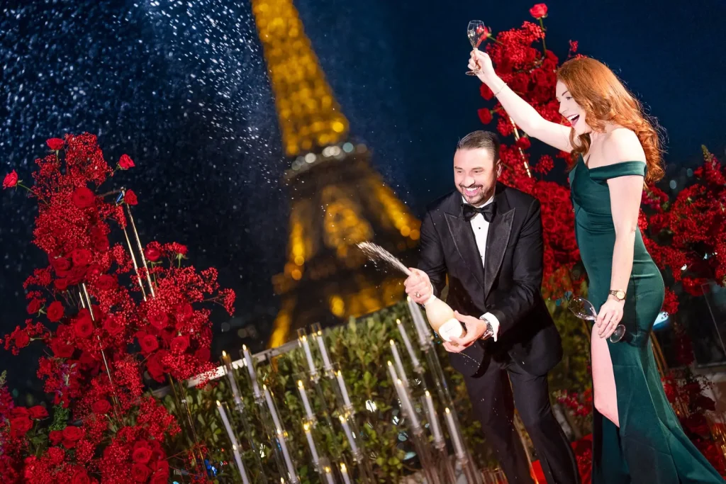 Joyful couple celebrating engagement by popping Champagne in front of a dramatic red floral backdrop at Shangri-La Hotel Paris, Eiffel Tower in view.