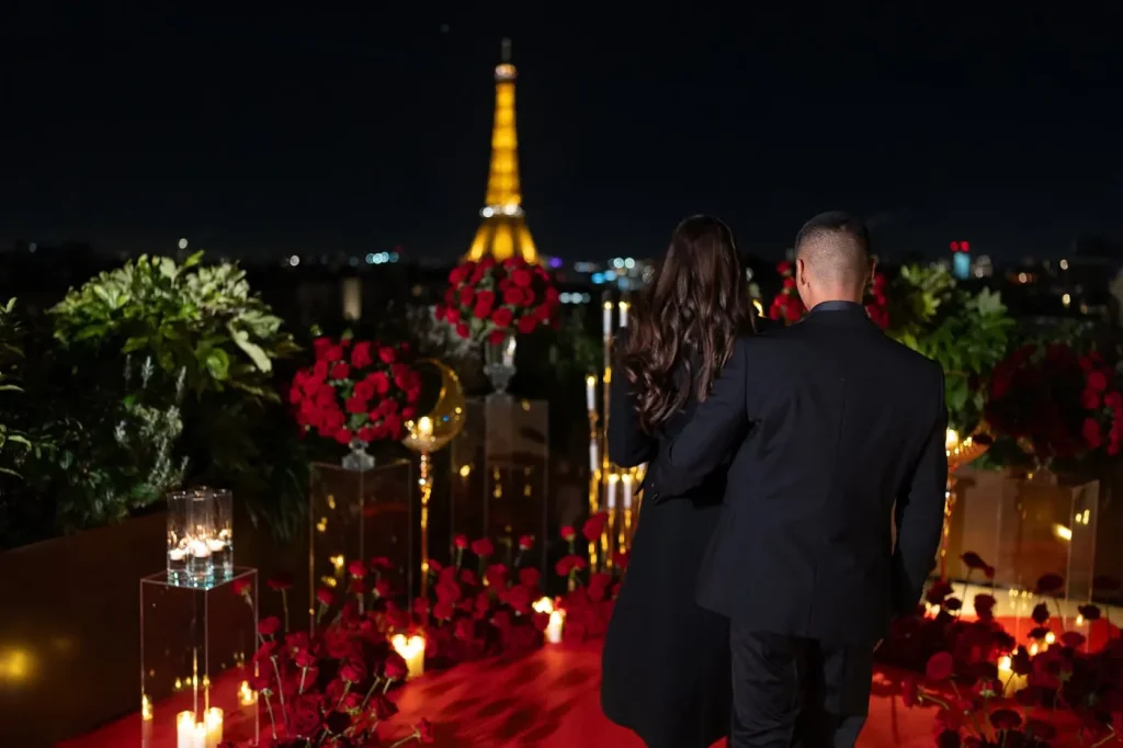 Ein Paar nähert sich nachts dem vorbereiteten Heiratsantrag auf der Dachterrasse des Peninsula Paris – rote Rosen, Kerzenlicht, Halbmonddekoration und der beleuchtete Eiffelturm vor ihnen.