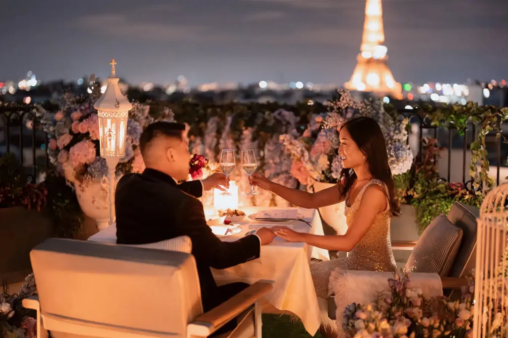 Ein frisch verlobtes Paar stößt mit Champagner bei einem Abendessen auf einer Pariser Dachterrasse im Kerzenschein an – umgeben von rosa Hortensien und Glyzinien, im Hintergrund der nachts beleuchtete Eiffelturm.