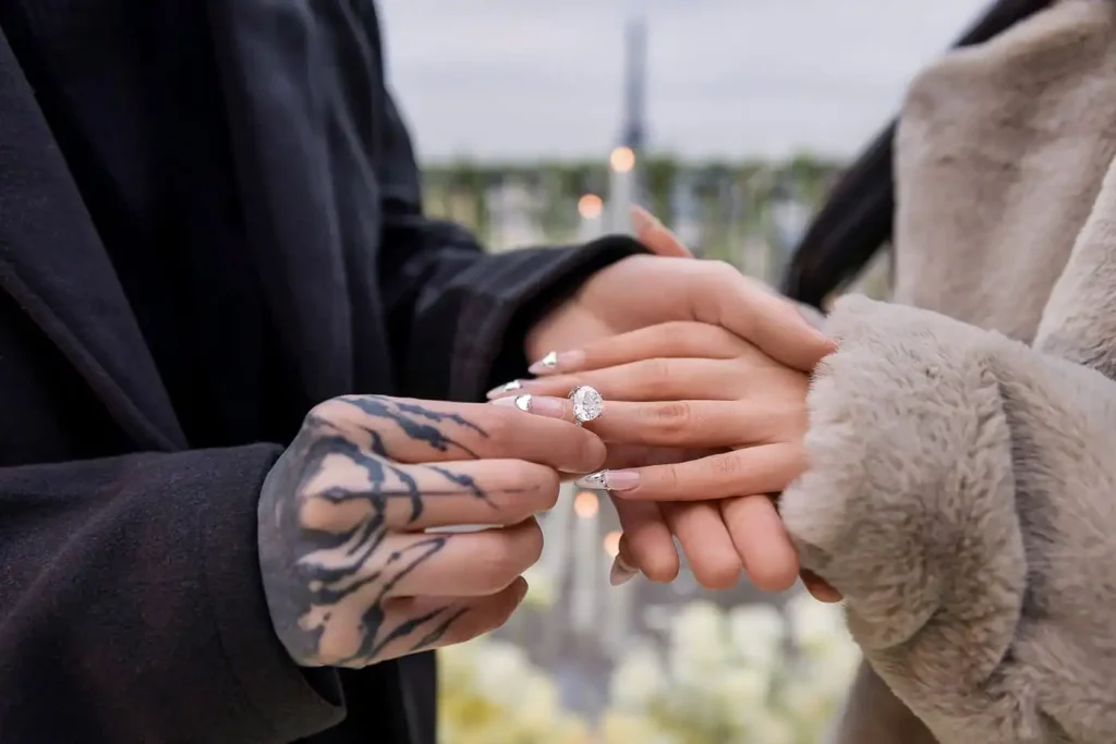 Nahaufnahme des Ringtauschs bei einem Heiratsantrag in Paris – eine tätowierte Hand schiebt einen birnenförmigen Diamant-Verlobungsring an ihren Ringfinger, der Eiffelturm ist im Hintergrund verschwommen zu sehen.