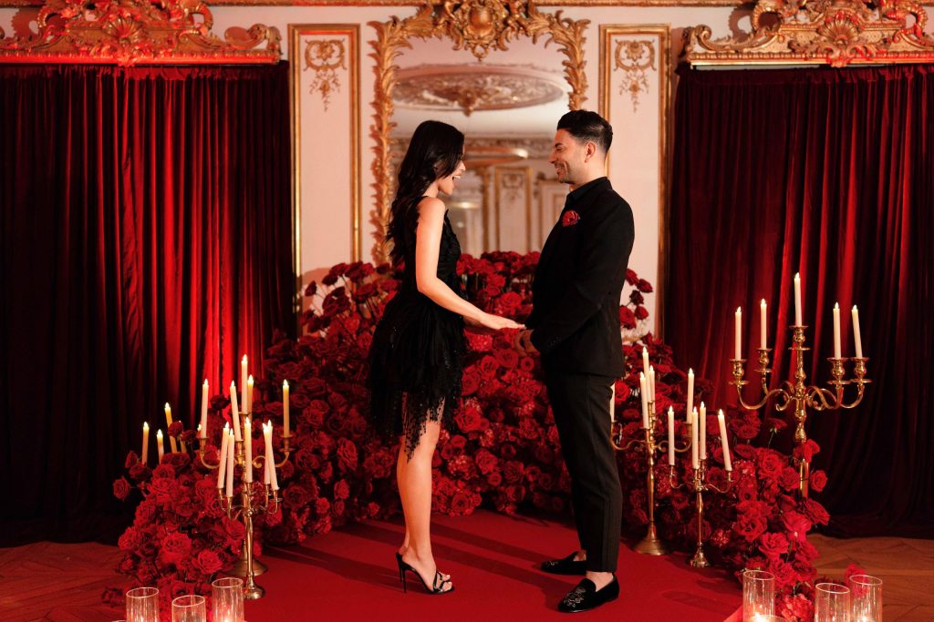 Heiratsantrag in einem Pariser Palastsalon – das Paar steht einander gegenüber, Hand in Hand; sie in einem schwarzen Tüll-Cocktailkleid lacht herzlich, er im schwarzen Anzug spricht. Umgeben ist der Raum von einer geschwungenen Installation aus roten Rosen, goldenen, mehrarmigen Kandelabern mit hohen, elfenbeinfarbenen Spitzen, schweren roten Samtvorhängen und einem vergoldeten Rokoko-Spiegel über einem Fischgrätparkettboden.