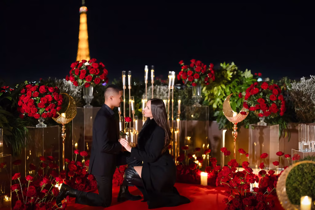Ein nächtlicher Heiratsantrag auf der Dachterrasse des Peninsula Paris Secret Table, der Eiffelturm in der Ferne beleuchtet, der Mann kniet mit einer einzelnen roten Rose, seine Partnerin kniet ebenfalls nieder, um ihm entgegenzukommen, umgeben von Hunderten roter Rosen, hohen Spitzkerzen, Halbmond-Dekoration und einem roten Teppich.