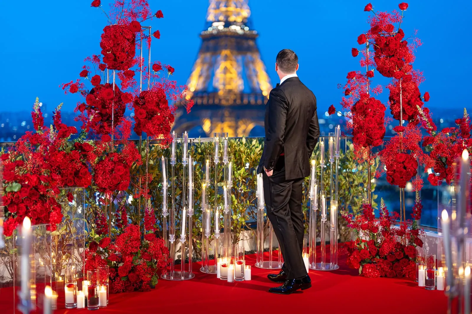 Le marié, en smoking, se trouve sur la terrasse du penthouse La Suite Shangri-La à l'heure bleue, face à la Tour Eiffel illuminée, au milieu d'installations de roses rouges haute couture et à la lueur des bougies, avant sa demande en mariage.